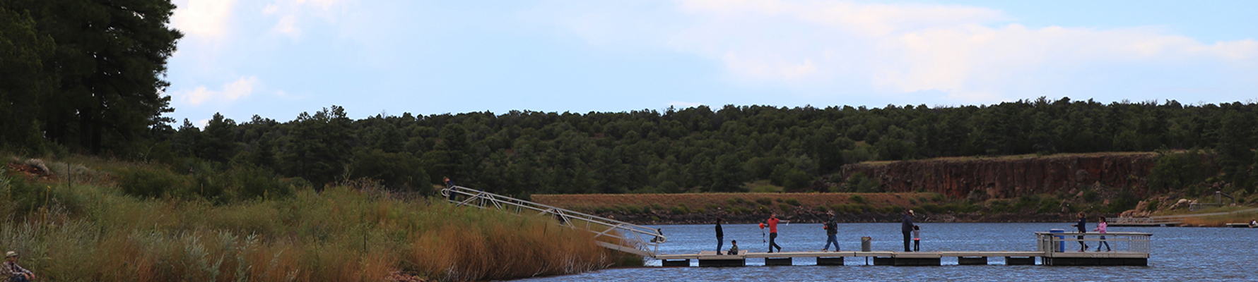 A group of anglers is fishing from a metal dock that floats in the ponderosa pine tree lined lake on a partly cloudy morning..