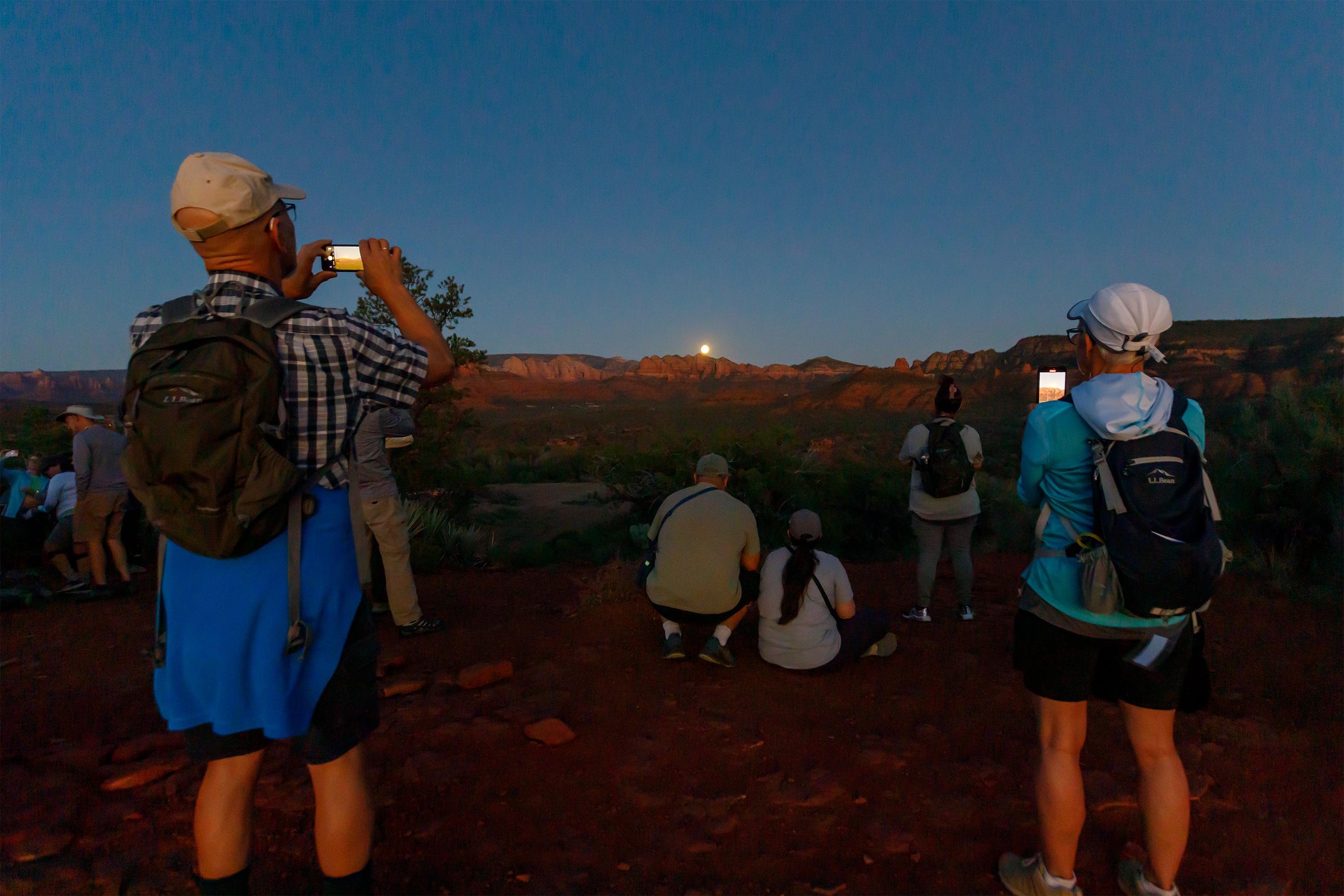 A group of hikers taking photos of the moon rising from behind a red rock escarpment in the distance.