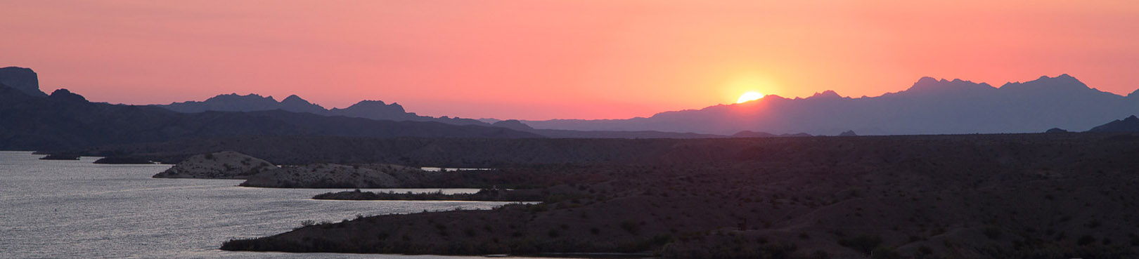 A sunset lights the sky orange as it sinks behind the mountains that border a lake.