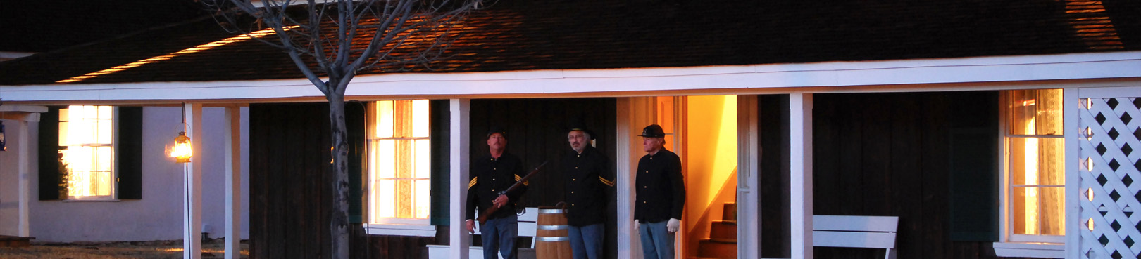Actors in period dress stand outside the historic Officer's Quarters at night. The house is illuminated with yellow-orange light.