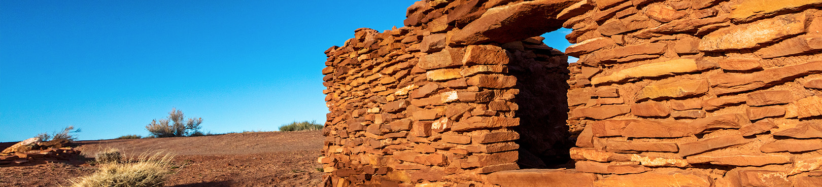 A stone sided pueblo replica in a desert landscape under a clear blue sky.