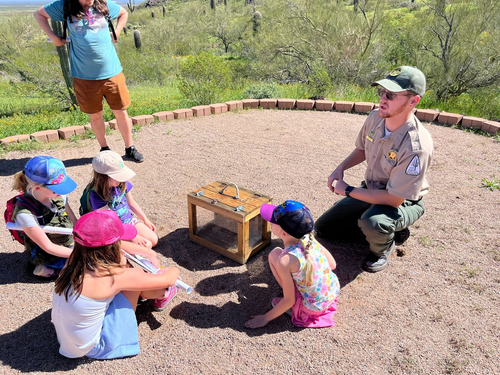 Children sit in a sandy area listening to a park ranger speak.
