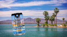 A photo of the greenish blue lake with a white sand beach and palm trees and mountains in the distance. A can of the Windsor Beach Blonde beer is overlaid on the photo.